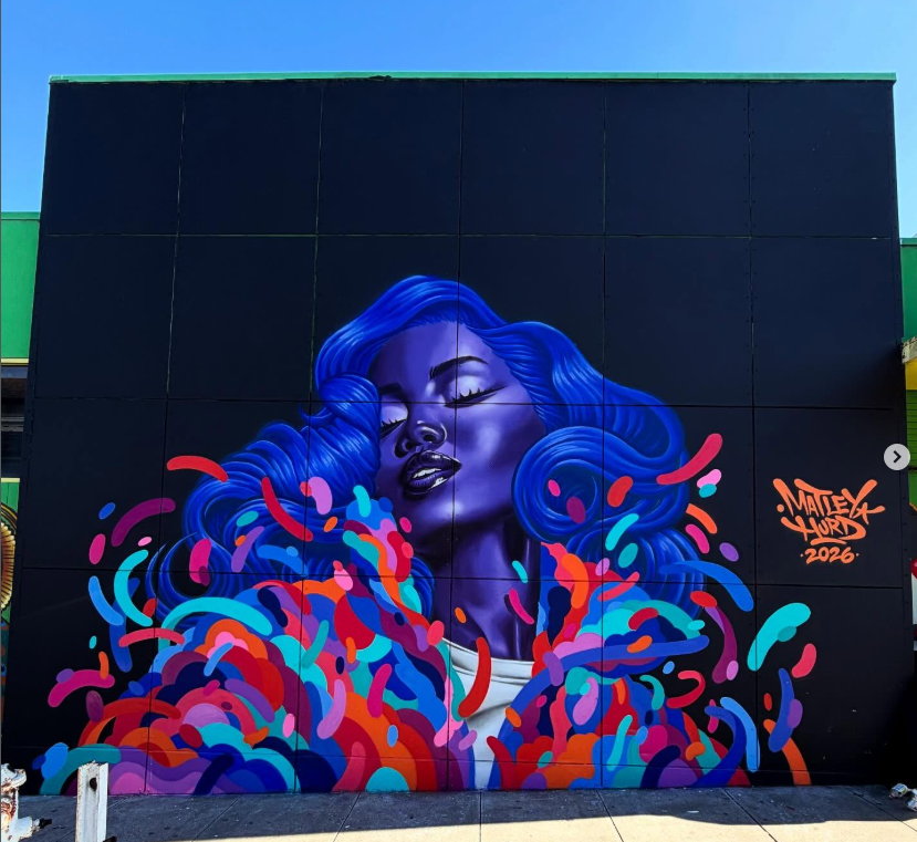 Vibrant wall mural by Matley Hurd on Bay Street San Franxcisco featuring a woman with blue hair and skin tone, surrounded by colorful flowing 3D-like ribbon patterns in red, coral, teal, and blue.