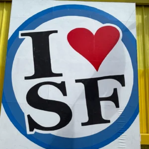 A square close-up shot of the "I Heart SF" mural logo, featuring a bold black "I", a red heart, and "SF" inside a white circle with a blue border on a yellow wall.