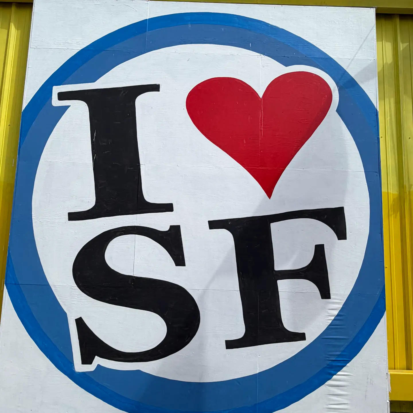 A square close-up shot of the "I Heart SF" mural logo, featuring a bold black "I", a red heart, and "SF" inside a white circle with a blue border on a yellow wall.