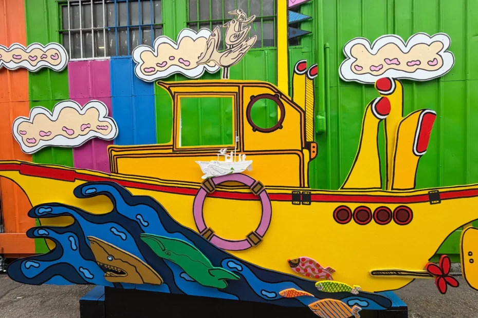 A fully painted, bright yellow cutout mural of a whimsical fishing boat riding blue waves with colorful fish, set against a vibrant corrugated wall at Umbrella Alley. The boat's nameplate area is left blank.