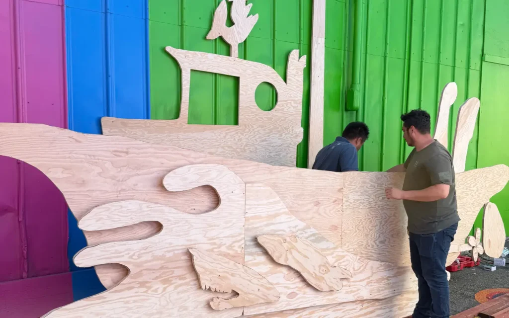 Two workers assembling a large, unpainted marine-grade plywood cutout of a Monterrey fishing boat against a bright pink, blue, and green wall at Umbrella Alle