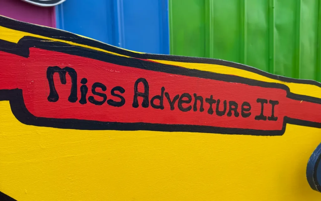 A close-up of the painted red banner reading "Miss Adventure II" in black lettering on the side of a bright yellow boat mural, set against a colorful blue, pink, and green corrugated background.