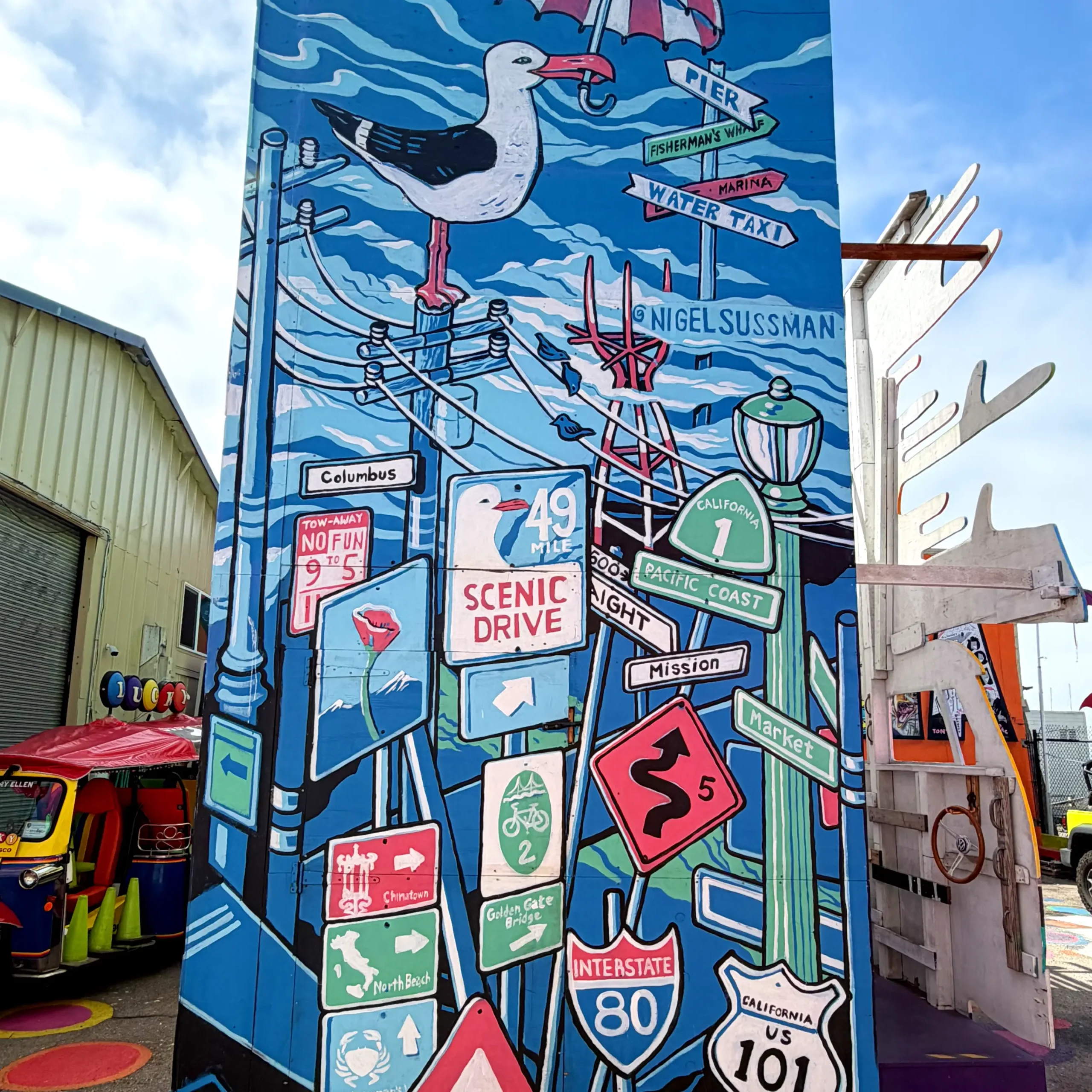 A panoramic view of Nigel Sussman’s vibrant street sign mural at Umbrella Alley. Painted on a freestanding pillar, the artwork features a chaotic stack of classic San Francisco signs, topped with a seagull, including the 49 Mile Scenic Drive and Highway 101.