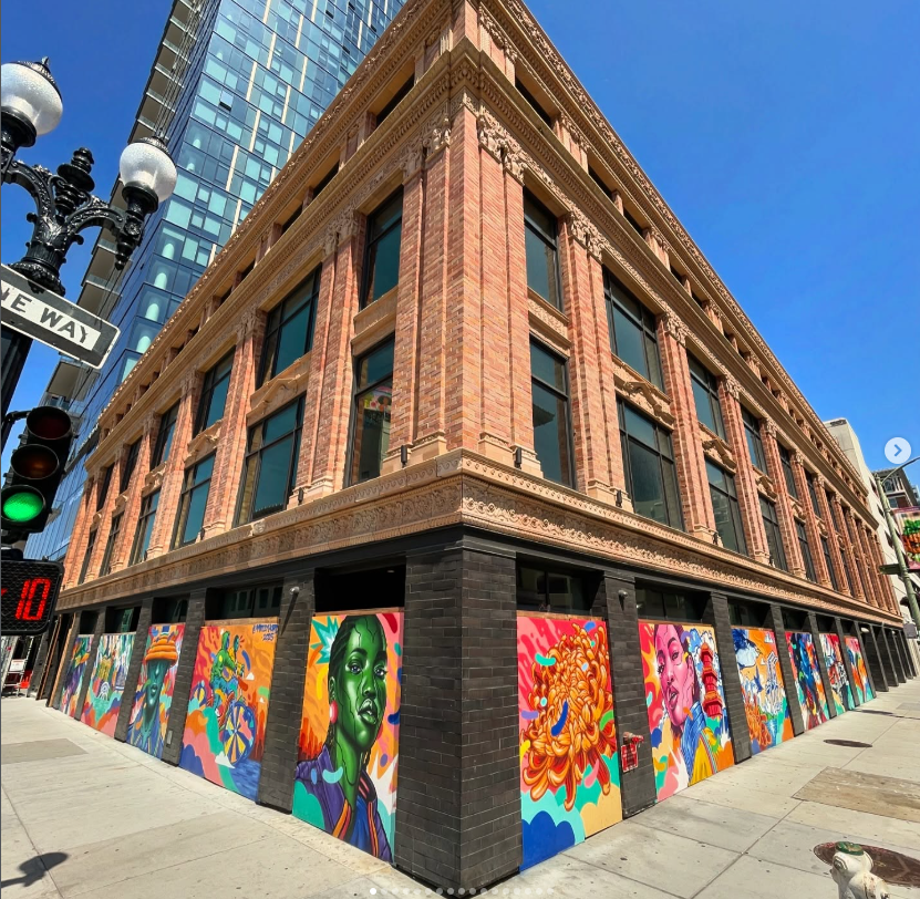 Street-level view of a classic brick building in downtown Oakland with ground-floor boarded windows painted with vibrant murals featuring colorful women's portraits, large flowers, and flowing abstract waves.