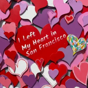 A square close-up of a mural showing overlapping painted hearts in shades of red, pink, and purple. A large red heart reads "I Left My Heart in San Francisco," with a brightly colored, patterned heart nestled on the right.