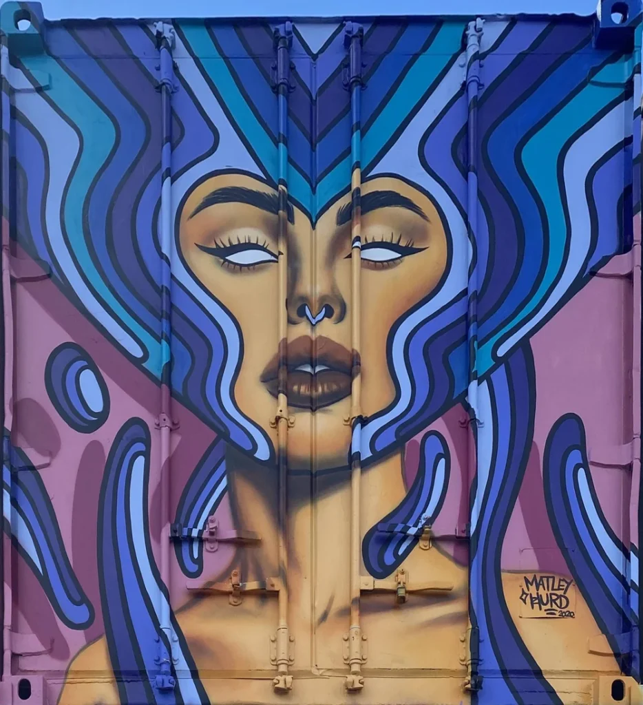 Mural of a Star Goddess with blue flowing hair, striking facial features, and glowing white eyes painted on a corrugated metal shipping container.